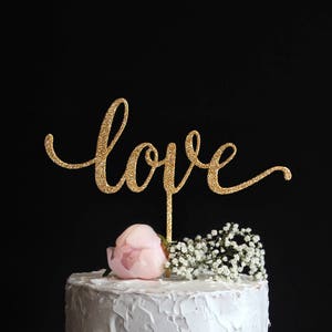 Op de afbeelding: Gouden glitter taarttopper met het woord "love" in cursief schrift. De topper staat op een witte taart met roze en witte bloemen.
