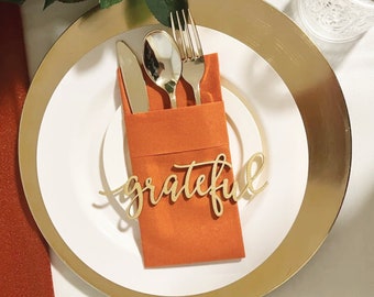 Könnte beinhalten: Ein weißer Teller mit goldenem Rand, eine gefaltete orangefarbene Serviette und goldenes Besteck. Die Serviette hat ein Holzschild mit der Aufschrift "grateful" in Kursivschrift.
