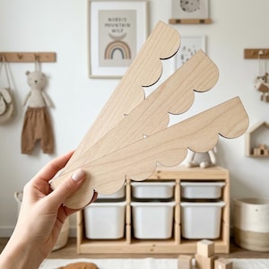 May include: Three light-colored wooden decorative pieces with a scalloped edge are held in a hand. The pieces are in a fan-like arrangement. The background includes a wooden storage unit with white bins, a framed picture, and children's decor.