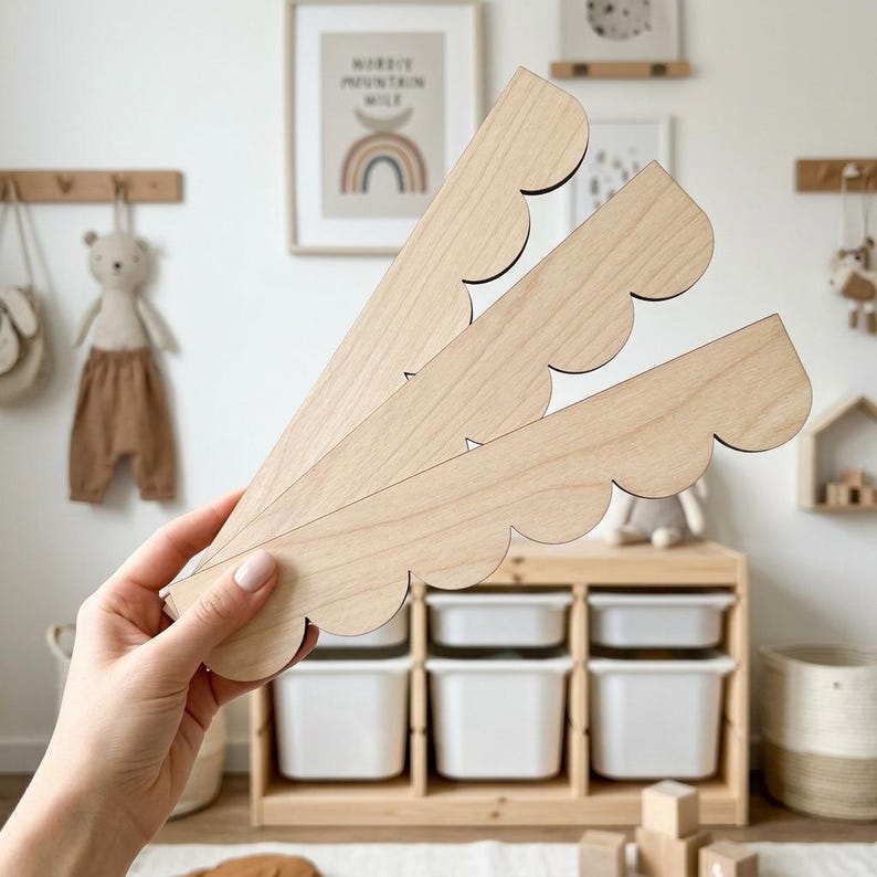 May include: Three light-colored wooden decorative pieces with a scalloped edge are held in a hand. The pieces are in a fan-like arrangement. The background includes a wooden storage unit with white bins, a framed picture, and children's decor.