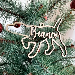 Op de afbeelding: Een houten kerstversiering in de vorm van een hond met de naam "Bianco" in cursief schrift. De versiering hangt aan een kerstboom met rode ballen. Het honden silhouet is lichtbruin.