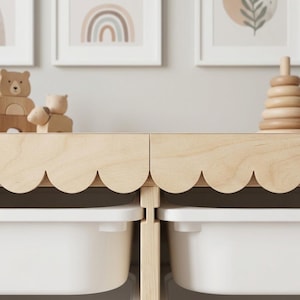 May include: A light wood storage unit with a scalloped edge design, featuring white storage bins. The unit is adorned with wooden toys and framed art prints with a rainbow and leaf theme, creating a nursery or playroom aesthetic.