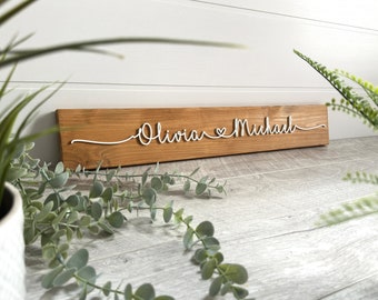 Puede incluir: Un letrero de madera con los nombres "Olivia" y "Michael" escritos en cursiva blanca con un símbolo de corazón entre los nombres.