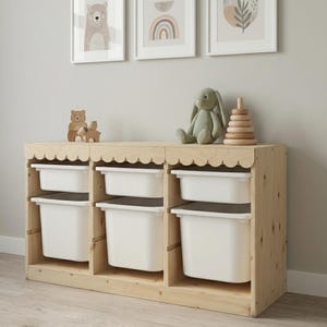 May include: A wooden storage unit with six white bins, topped with a scalloped edge. The unit is made of light-colored wood and has a natural finish. Decorative items, including a wooden bear, a stuffed bunny, and a wooden stacking toy, sit on top.