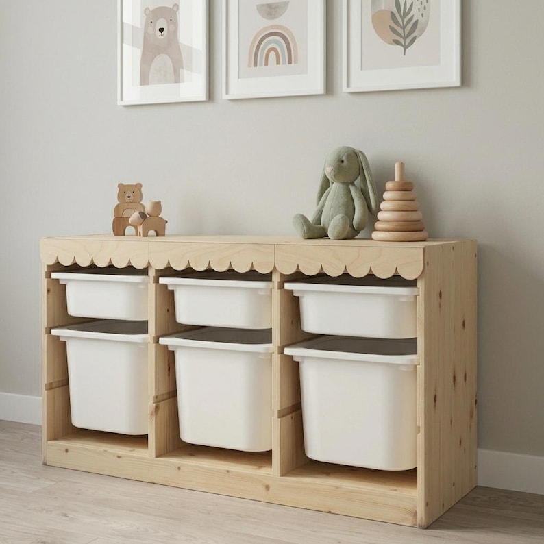 May include: A wooden storage unit with six white bins, topped with a scalloped edge. The unit is made of light-colored wood and has a natural finish. Decorative items, including a wooden bear, a stuffed bunny, and a wooden stacking toy, sit on top.
