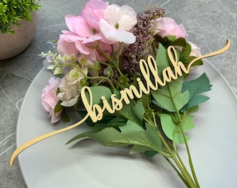 Op de afbeelding: Een gouden houten bord met het woord "bismillah" in cursief schrift. Het bord ligt op een wit bord met een boeket roze en witte bloemen.