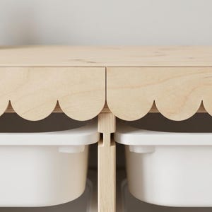 May include: A light-colored wooden storage unit with a scalloped edge design. The unit has two white storage bins visible below. The wood has a natural finish, and the bins are a solid white color. The design is simple and modern.