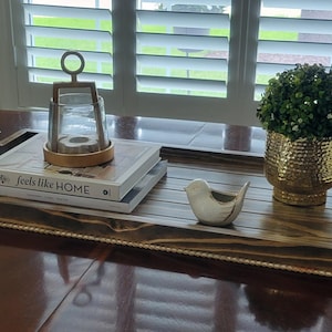 May include: A decorative wooden tray with black handles holds a stack of books, a glass lantern, and a small ceramic bird. A gold-colored textured vase with a green plant sits to the right. The tray is on a dark brown table.