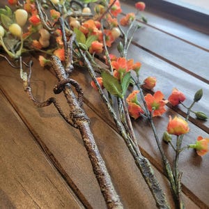 May include: Close-up of artificial floral stems with orange and yellow blossoms, green leaves, and small white berries. The stems have a textured, brown appearance, and are arranged on a wooden surface.