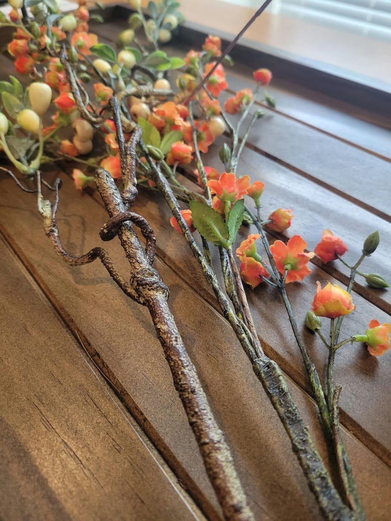 May include: Close-up of artificial floral stems with orange and yellow blossoms, green leaves, and small white berries. The stems have a textured, brown appearance, and are arranged on a wooden surface.