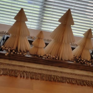May include: A wooden tray with four white paper Christmas trees decorated with gold glitter. The tray is sitting on a beige table runner with a fringe border.