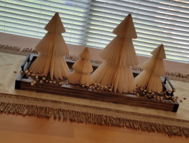 May include: A wooden tray with four white paper Christmas trees decorated with gold glitter. The tray is sitting on a beige table runner with a fringe border.