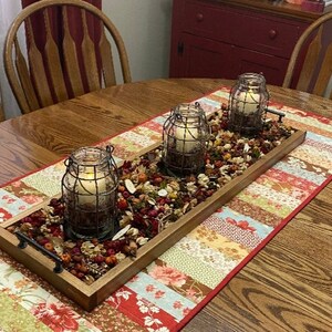 May include: A wooden tray with three glass candle holders with wire cages, filled with dried flowers and leaves, on a colorful patchwork table runner.