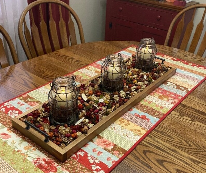 May include: A wooden tray with three glass candle holders with wire cages, filled with dried flowers and leaves, on a colorful patchwork table runner.