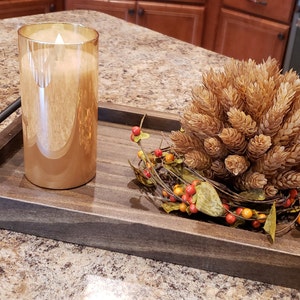 May include: A wooden tray with a black handle, a gold candle, and a fall-themed centerpiece with dried brown pinecones and red berries.