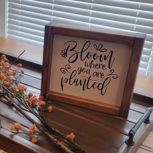 May include: A wooden framed sign with the quote "Bloom where you are planted" in black script. The sign is on a wooden tray with a floral arrangement of orange flowers and brown branches.