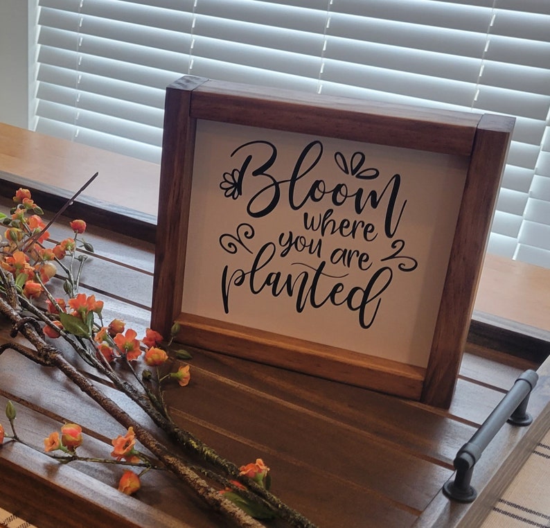 May include: A wooden framed sign with the quote "Bloom where you are planted" in black script. The sign is on a wooden tray with a floral arrangement of orange flowers and brown branches.