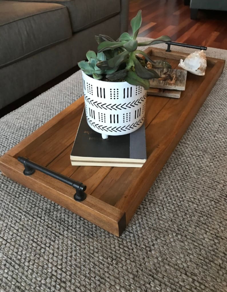 May include: A wooden tray with a black metal handle, a white planter with a black geometric pattern, a book, and a seashell. The tray is sitting on a gray rug.