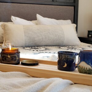 May include: A wooden tray with a lit candle, a jar of candle wax, and two blue ceramic mugs with moon and mountain designs. The tray is on a bed with a white blanket and pillows.