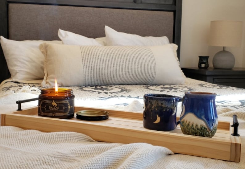 May include: A wooden tray with a lit candle, a jar of candle wax, and two blue ceramic mugs with moon and mountain designs. The tray is on a bed with a white blanket and pillows.