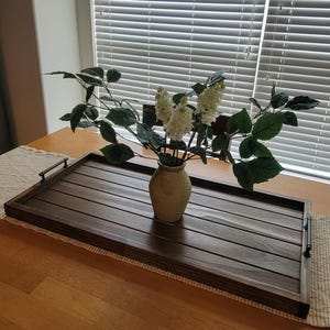 May include: A rectangular wooden serving tray with dark brown slats and black handles. A beige vase holds white flowers and green leaves. The tray is on a wooden table with a beige runner.