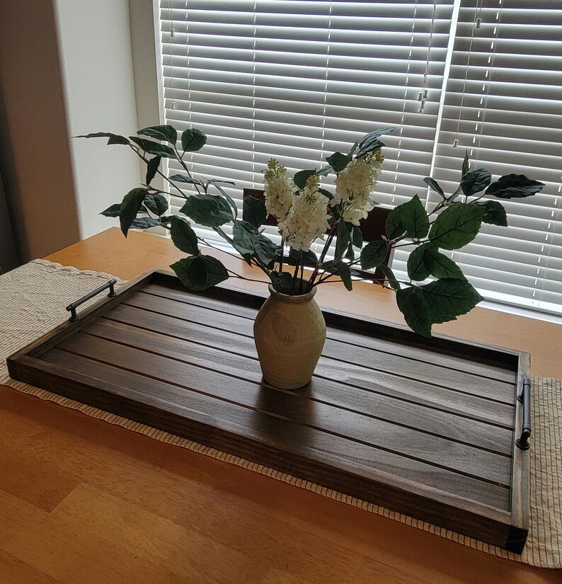 May include: A rectangular wooden serving tray with dark brown slats and black handles. A beige vase holds white flowers and green leaves. The tray is on a wooden table with a beige runner.