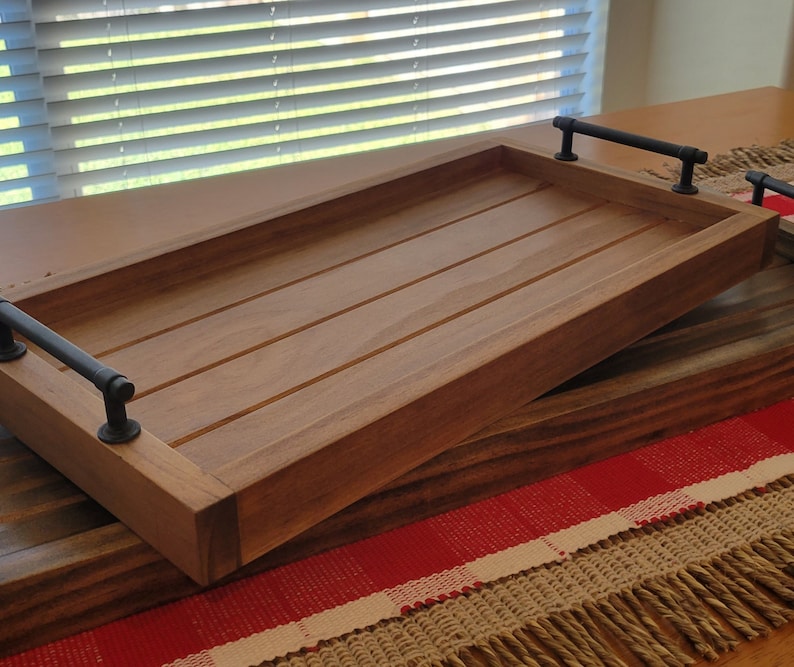 May include: A wooden serving tray with black metal handles. The tray has a natural wood finish and is made of solid wood. The tray is sitting on a red and white checkered rug.