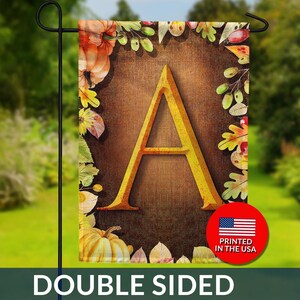 May include: Double-sided garden flag featuring a large gold letter "A" on a brown background, surrounded by autumn leaves and pumpkins. The flag has a rustic, fall theme and includes the text "DOUBLE SIDED". A red circle with the text "PRINTED IN THE USA" is also visible.