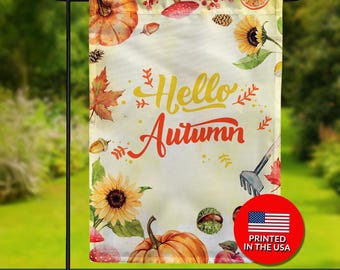 May include: Double-sided garden flag with a watercolor design featuring pumpkins, sunflowers, and autumn leaves. The flag has the words "Hello Autumn" in yellow and orange. A red circle with the US flag and "Printed in the USA" is visible.