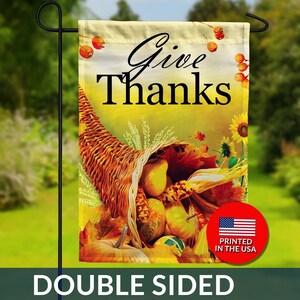 May include: A double-sided garden flag with a Thanksgiving theme. The flag features a cornucopia overflowing with pumpkins, gourds, and fall foliage. The words "Give Thanks" are written in elegant script. A red circle with the American flag and "Printed in the USA" is visible.