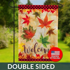 May include: Double-sided garden flag with a fall theme. The flag features colorful maple leaves in shades of red, orange, yellow, and green against a textured background. The word "Welcome" is written in a cursive font. A red and black plaid border accents the top and bottom.