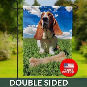 May include: Double-sided garden flag featuring a Basset Hound sitting in green grass with a large bone. The background shows a blue sky with white clouds. The flag has a red circle with the American flag and the text "PRINTED IN THE USA".