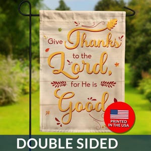 May include: A double-sided garden flag with the message "Give Thanks to the Lord, for He is Good" in gold script. The flag has a light beige background with a wood-like texture and decorative fall-themed accents. A red circle with the American flag and "Printed in the USA" is on the bottom right.