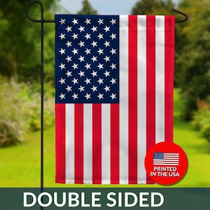 May include: A double-sided garden flag featuring the American flag design. The flag has a blue field with white stars and red and white stripes. A red circle with the text "PRINTED IN THE USA" is visible. The flag is hanging on a black metal pole.