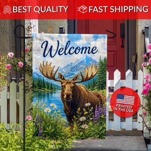 May include: A rectangular garden flag featuring a moose standing in a field of wildflowers near a blue lake with a mountain range in the background. The word Welcome is printed in dark blue script at the top of the flag above the moose.