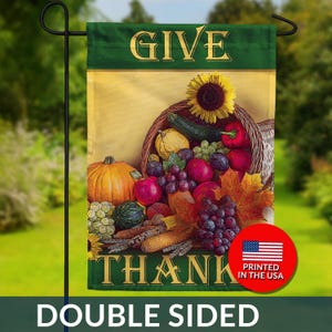 May include: Double-sided garden flag with a Thanksgiving theme. The flag features a cornucopia overflowing with fruits and vegetables, with the words "GIVE" and "THANK" in gold lettering. A sunflower and the text "PRINTED IN THE USA" are also visible.