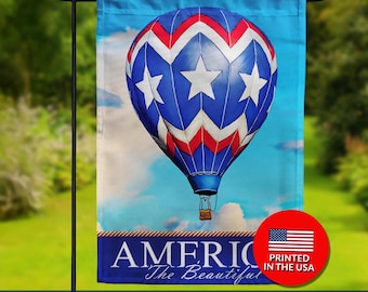May include: A double-sided garden flag featuring a patriotic hot air balloon design. The balloon is blue with white stars and red accents, set against a sky background. The flag includes the text "AMERICA The Beautiful" and "PRINTED IN THE USA".