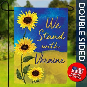 May include: Double-sided garden flag with sunflowers and the text "We Stand with Ukraine" in yellow. The flag features a blue and yellow background, with the words printed in a stylized font. A red circle with the American flag and "Printed in the USA" is in the bottom right corner.