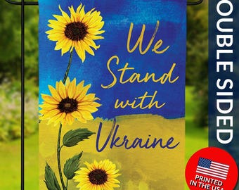 May include: Double-sided garden flag with sunflowers and the text "We Stand with Ukraine" in yellow. The flag features a blue and yellow background, with the words printed in a stylized font. A red circle with the American flag and "Printed in the USA" is in the bottom right corner.