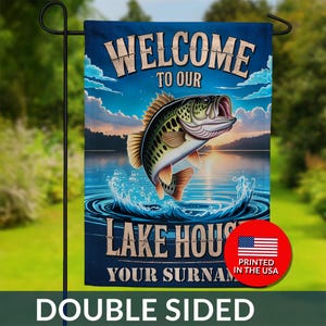 May include: Double-sided garden flag with a lake house theme. The flag features a leaping bass fish illustration above the text "WELCOME TO OUR LAKE HOUSE". The background shows a sunset over a lake. The flag is printed in the USA.