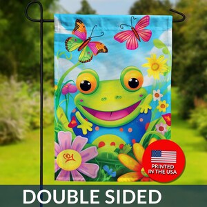 May include: Double-sided garden flag featuring a cheerful frog with a big smile, surrounded by colorful flowers and butterflies against a blue sky. The flag has the text "DOUBLE SIDED" and a "PRINTED IN THE USA" logo.