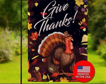 May include: A double-sided garden flag with a Thanksgiving theme. The flag features a cartoon turkey wearing a hat, surrounded by autumn leaves, and the words "Give Thanks!". A red circle with the American flag and "Printed in the USA" is visible.