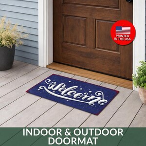May include: A rectangular blue doormat with the word "Welcome" in white script. The mat has a red border and white speckles. The words "INDOOR & OUTDOOR DOORMAT" are at the bottom. A red circle with the American flag and "PRINTED IN THE USA" is visible.