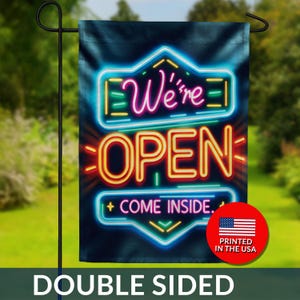May include: Double-sided garden flag with a neon sign design. The flag features the words "We're OPEN" and "+ COME INSIDE." in bright colors against a dark background. A red circle with the American flag and "PRINTED IN THE USA" is in the bottom right corner.