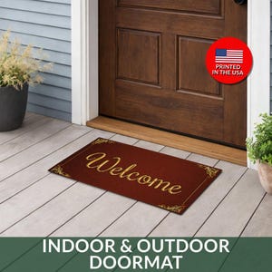 May include: A rectangular, burgundy doormat with the word "Welcome" in gold script. The mat has a textured appearance and gold decorative corners. The image also includes the text "INDOOR & OUTDOOR DOORMAT" and a circular graphic that says "PRINTED IN THE USA".