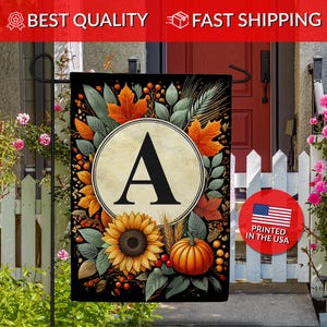 May include: A decorative garden flag featuring a large letter "A" in a cream-colored circle, surrounded by autumn leaves, a sunflower, and a pumpkin. The flag is printed in the USA and is displayed on a black metal stand.