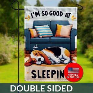 May include: Double-sided garden flag featuring a cartoon dog sleeping in front of a blue couch with pillows. The flag reads "I'M SO GOOD AT SLEEPING" with a USA flag graphic. The flag is designed for outdoor use.