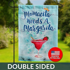 May include: A double-sided garden flag with the text "Mamacita Needs A Margarita" in blue script. The flag features a watercolor illustration of a margarita with a strawberry garnish. A small American flag graphic is in the bottom right corner.