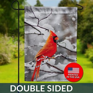 May include: A double-sided garden flag featuring a vibrant red cardinal perched on a snow-covered branch. The background is a grayscale winter scene. The flag has the text "DOUBLE SIDED" and a "PRINTED IN THE USA" logo.