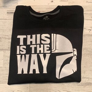 Puede incluir: Camiseta negra con un estampado gr&aacute;fico blanco de un casco de Mandaloriano y el texto "This is the way".